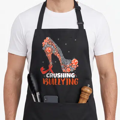 Prevention unity day apron - Diamond high heel shoe with red ribbon and crushing bullying text — shown on a black cotton canvas apron against a white background