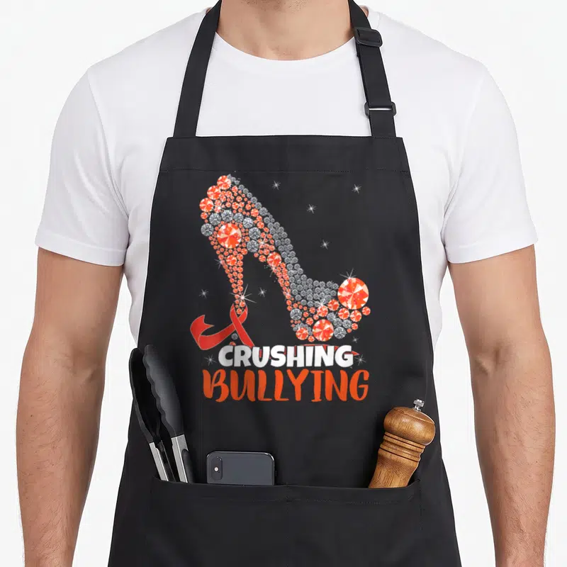 Prevention unity day apron - Diamond high heel shoe with red ribbon and crushing bullying text — shown on a black cotton canvas apron against a white background