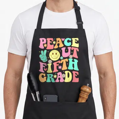 Peace groovy grade apron - Stacked retro text PEACE OUT FIFTH GRADE with peace sign, smiley face, and flowers in pink — shown on a black cotton canvas apron against a white background