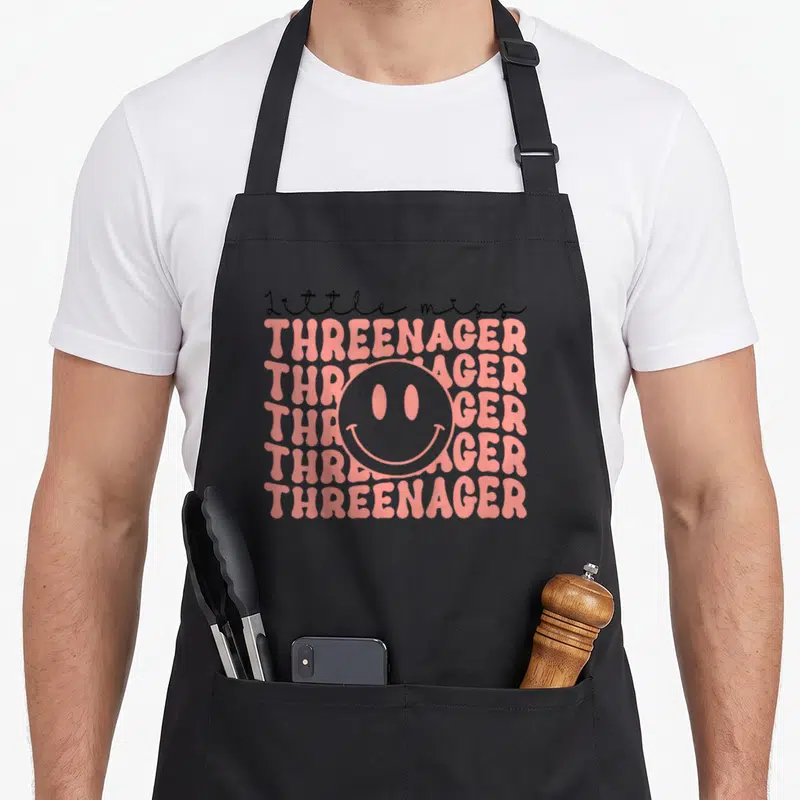 Threenager boho apron - Pink stacked text reading threenager with black script and smiley face — shown on a black cotton canvas apron against a white background