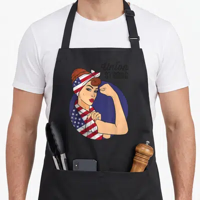 Union rosie the riveter apron - Woman flexing arm wearing US flag bandana with Union STRONG text on blue circle — shown on a black cotton canvas apron against a white background