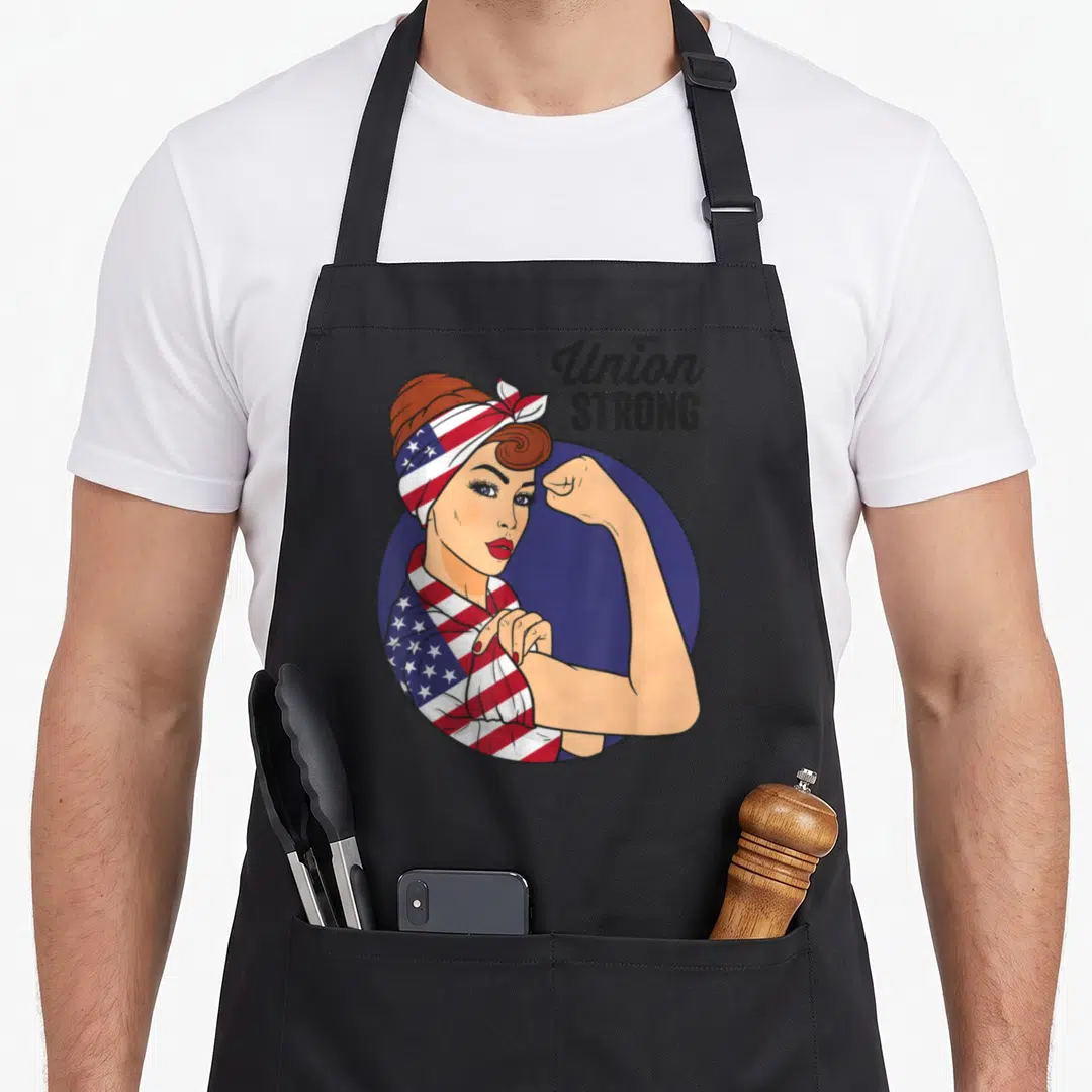 Union rosie the riveter apron - Woman flexing arm wearing US flag bandana with Union STRONG text on blue circle — shown on a black cotton canvas apron against a white background