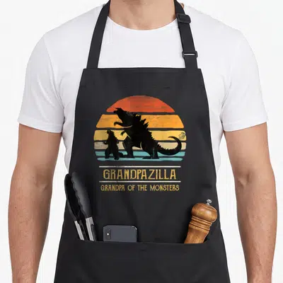 Grandpa funny halloween apron - Two black Godzilla silhouettes over a striped retro sun with Grandpapazilla text — shown on a black cotton canvas apron against a white background