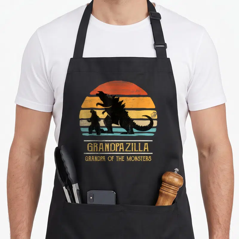 Grandpa funny halloween apron - Two black Godzilla silhouettes over a striped retro sun with Grandpapazilla text — shown on a black cotton canvas apron against a white background