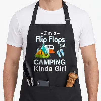 Retro camping apron - I'm a Flip Flops and CAMPING Kinda Girl text with camper tent and flip flops illustration — shown on a black cotton canvas apron against a white background