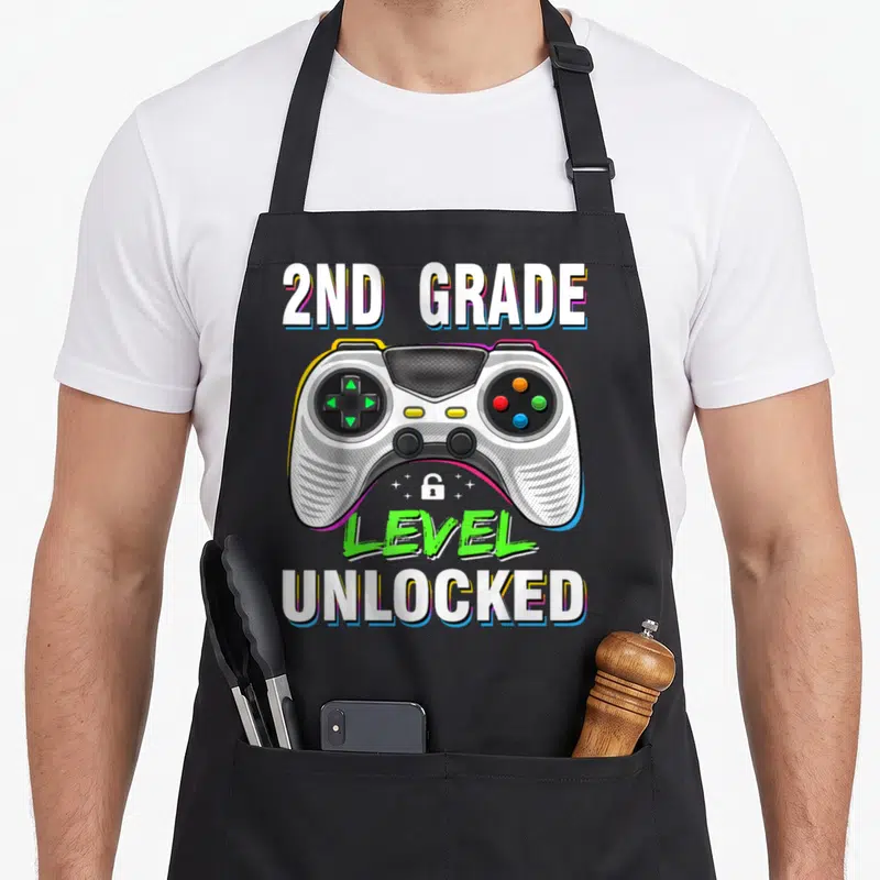 Level 2nd apron - 2ND GRADE LEVEL UNLOCKED text with game controller and small lock icon in white grey green blue — shown on a black cotton canvas apron against a white background