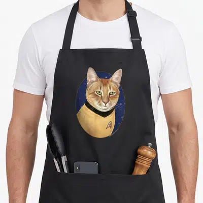 Captain cat apron - Realistic orange cat in yellow uniform inside dark blue oval with white stars — shown on a black cotton canvas apron against a white background