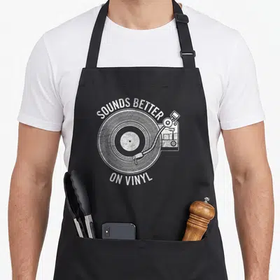 Better vinyl apron - White distressed record player graphic with arched text SOUNDS BETTER ON VINYL — shown on a black cotton canvas apron against a white background