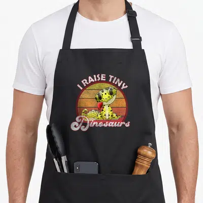 Raise tiny leopard apron - Cartoon gecko on retro striped sun with I RAISE TINY Dinosaurs text — shown on a black cotton canvas apron against a white background