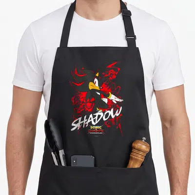 Shadow x apron - Shadow the Hedgehog cartoon figure with red splatters and white SHADOW script lettering — shown on a black cotton canvas apron against a white background