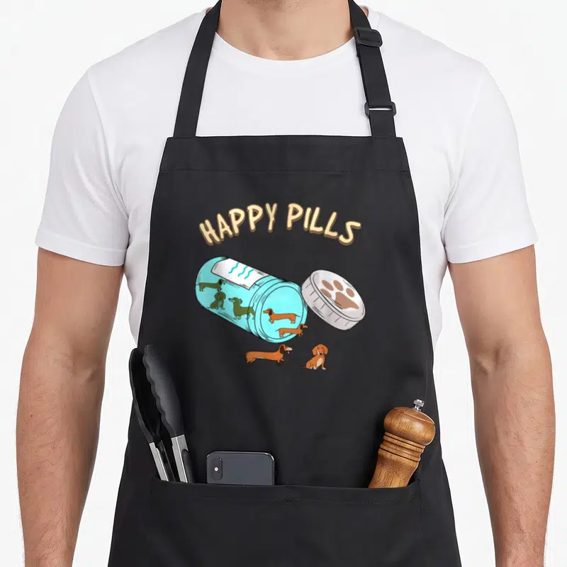 Pill dachshund apron - HAPPY PILLS text over blue bottle with brown dachshunds and paw print cap — shown on a black cotton canvas apron against a white background