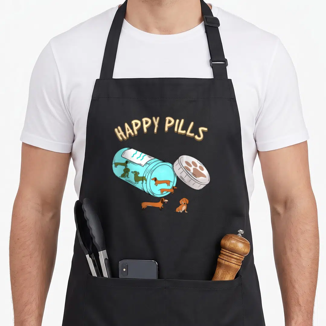 Pill dachshund apron - HAPPY PILLS text over blue bottle with brown dachshunds and paw print cap — shown on a black cotton canvas apron against a white background