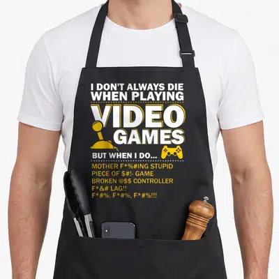 Playing gaming apron - White and yellow text reading I DON'T ALWAYS DIE with joystick and gamepad icons — shown on a black cotton canvas apron against a white background
