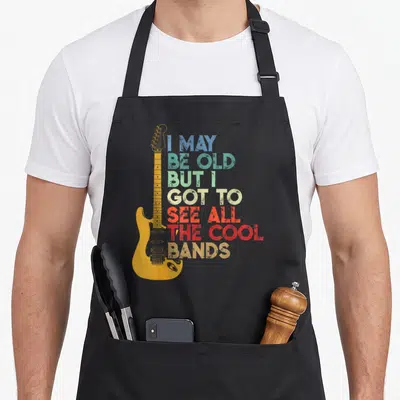 But may apron - Yellow electric guitar beside stacked multi-colored distressed text reading I MAY BE OLD BUT I GOT — shown on a black cotton canvas apron against a white background