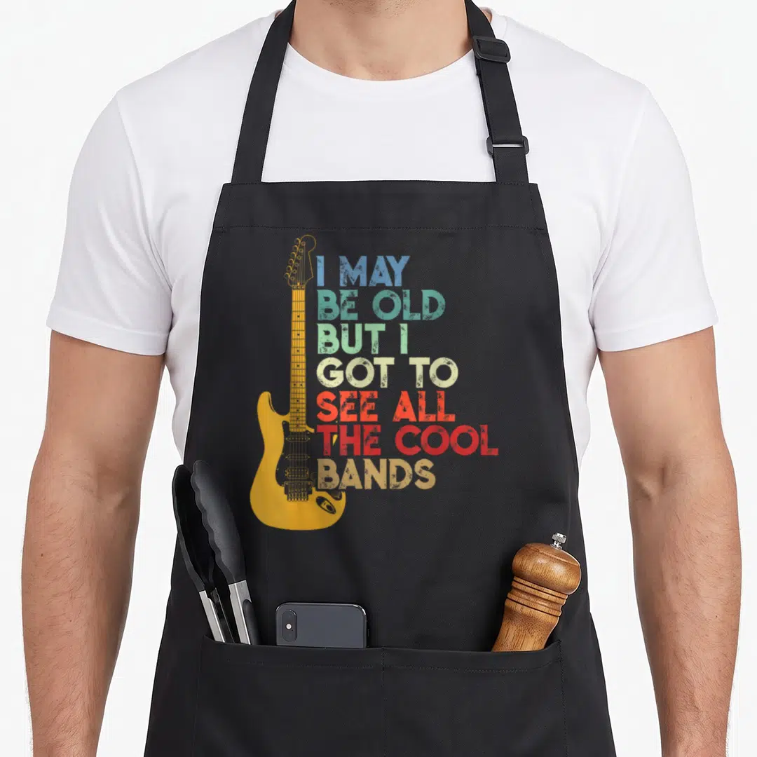 But may apron - Yellow electric guitar beside stacked multi-colored distressed text reading I MAY BE OLD BUT I GOT — shown on a black cotton canvas apron against a white background