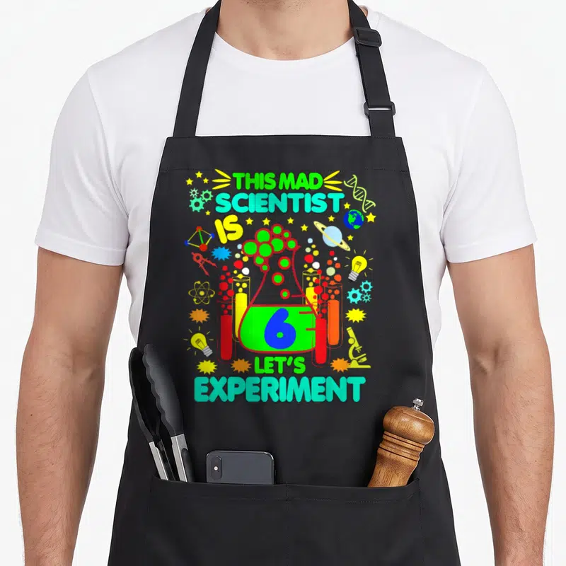 Birthday age science apron - Cartoon beaker with number 6 and science symbols like gears and DNA in green blue yellow — shown on a black cotton canvas apron against a white background