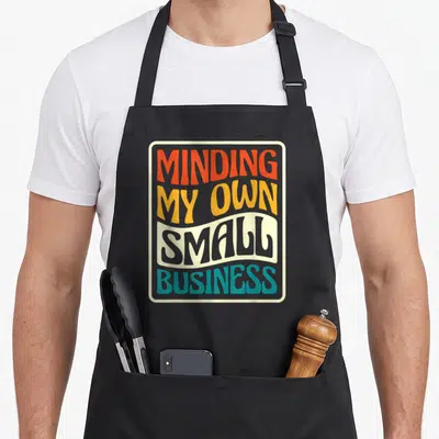 Minding owner small business aprons - Stacked MINDING MY OWN SMALL BUSINESS text in orange yellow grey teal beige — shown on a black cotton canvas apron against a white background