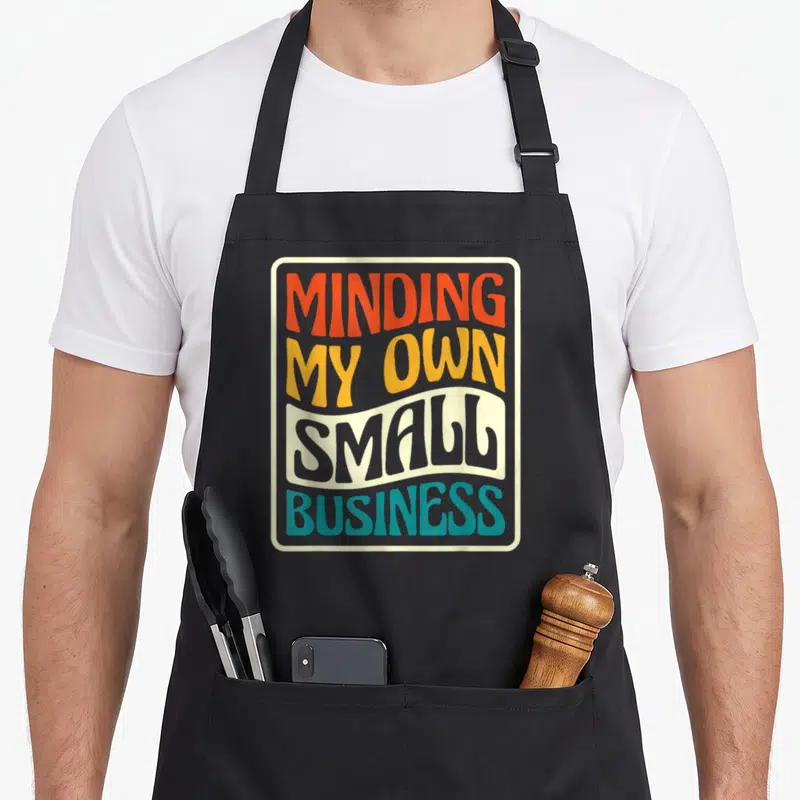 Minding owner small business aprons - Stacked MINDING MY OWN SMALL BUSINESS text in orange yellow grey teal beige — shown on a black cotton canvas apron against a white background
