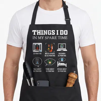 Spare printers apron - Cartoon 3D printer grid with text listing spare time activities in white and orange — shown on a black cotton canvas apron against a white background