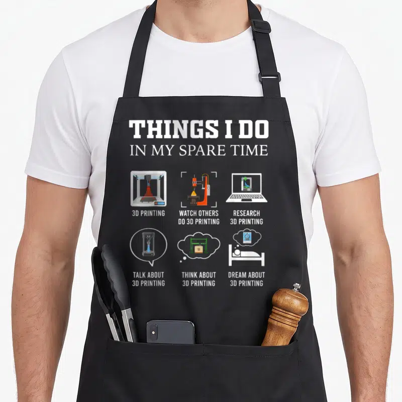 Spare printers apron - Cartoon 3D printer grid with text listing spare time activities in white and orange — shown on a black cotton canvas apron against a white background