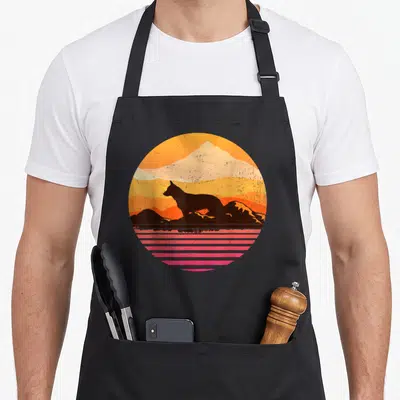 Heeler pet apron - Retro circular print of black dog silhouette, orange mountains, and pink horizontal lines — shown on a black cotton canvas apron against a white background