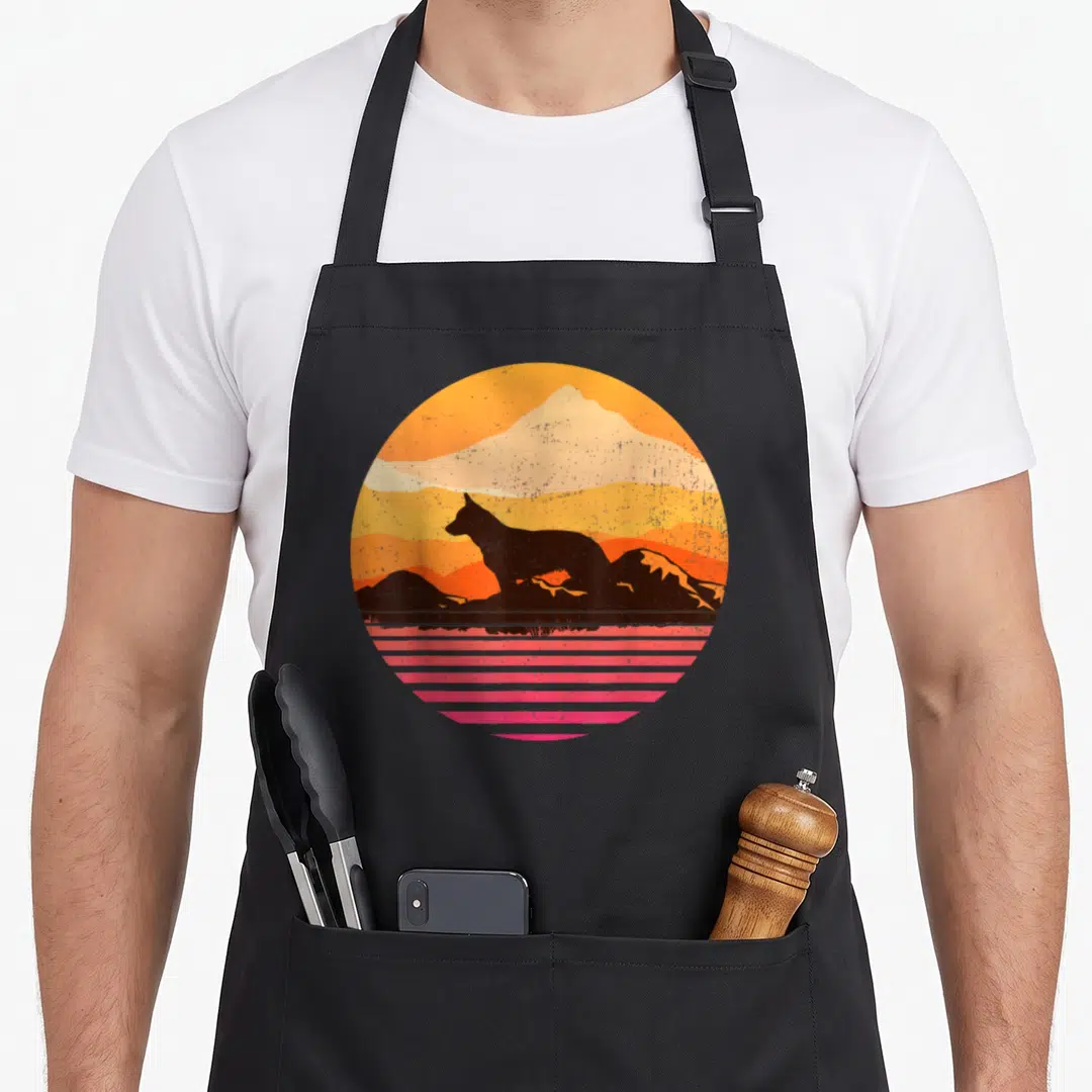 Heeler pet apron - Retro circular print of black dog silhouette, orange mountains, and pink horizontal lines — shown on a black cotton canvas apron against a white background