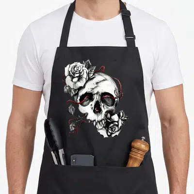 Skull rose apron - Line art skull with two white roses and red thorns piercing eye sockets — shown on a black cotton canvas apron against a white background