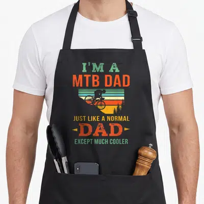 Biking biker bike apron - Retro biker silhouette against striped sunset with stacked MTB Dad text in orange yellow — shown on a black cotton canvas apron against a white background