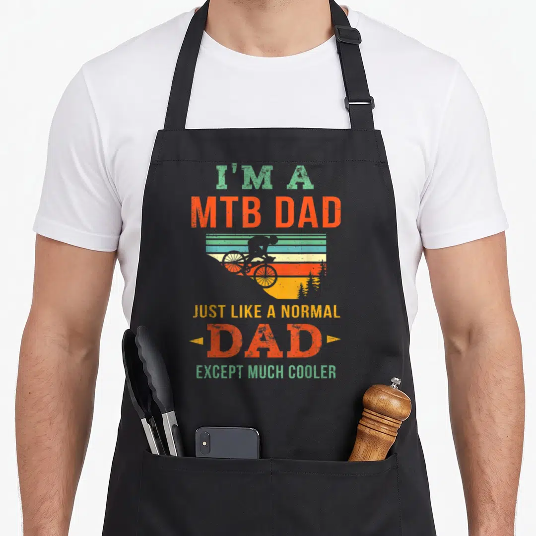 Biking biker bike apron - Retro biker silhouette against striped sunset with stacked MTB Dad text in orange yellow — shown on a black cotton canvas apron against a white background
