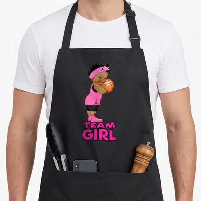 Ethnic gender reveal apron - Cartoon baby girl holding orange basketball with pink TEAM GIRL text below — shown on a black cotton canvas apron against a white background