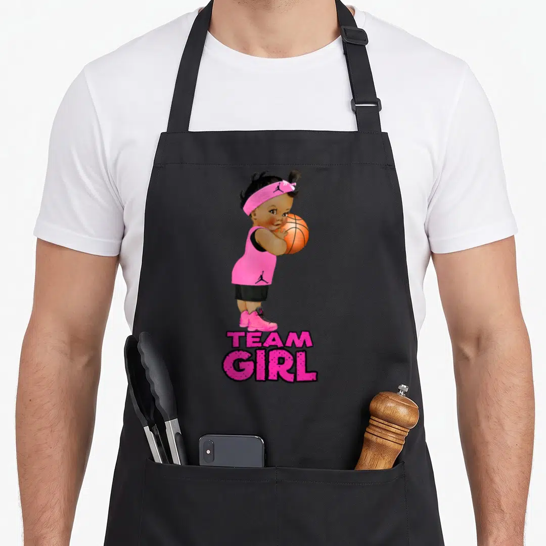 Ethnic gender reveal apron - Cartoon baby girl holding orange basketball with pink TEAM GIRL text below — shown on a black cotton canvas apron against a white background