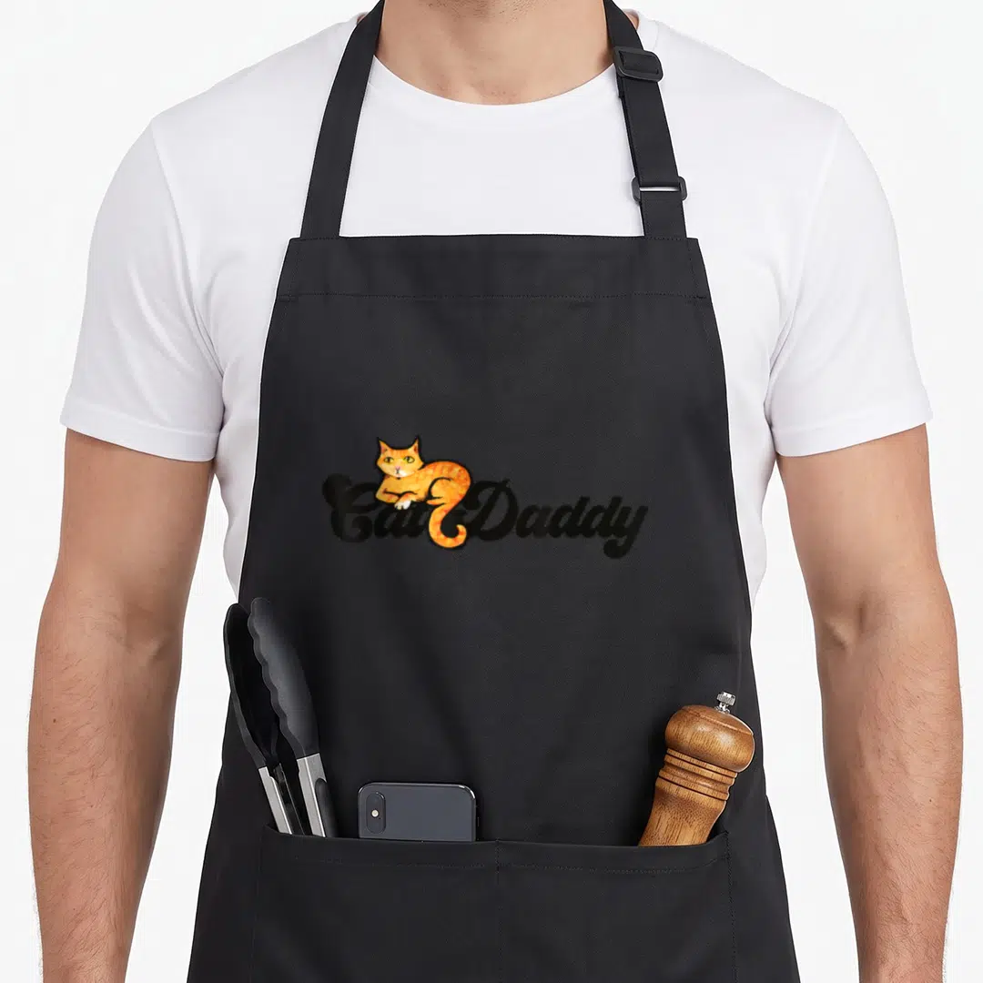 Humour cat dad apron - Orange cartoon cat resting on black Cat Daddy cursive text — shown on a black cotton canvas apron against a white background