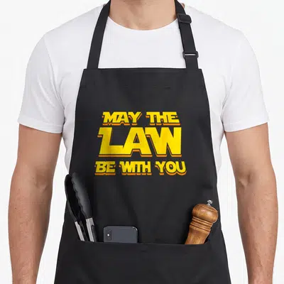 May lawyer apron - Stacked yellow text MAY THE LAW BE WITH YOU with orange outlines — shown on a black cotton canvas apron against a white background