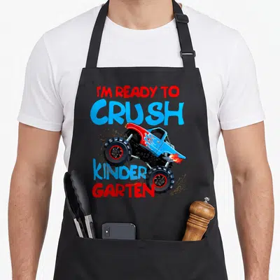 School kindergarten truck apron - Monster truck graphic with I'M READY TO CRUSH KINDER GARTEN text in red and blue — shown on a black cotton canvas apron against a white background