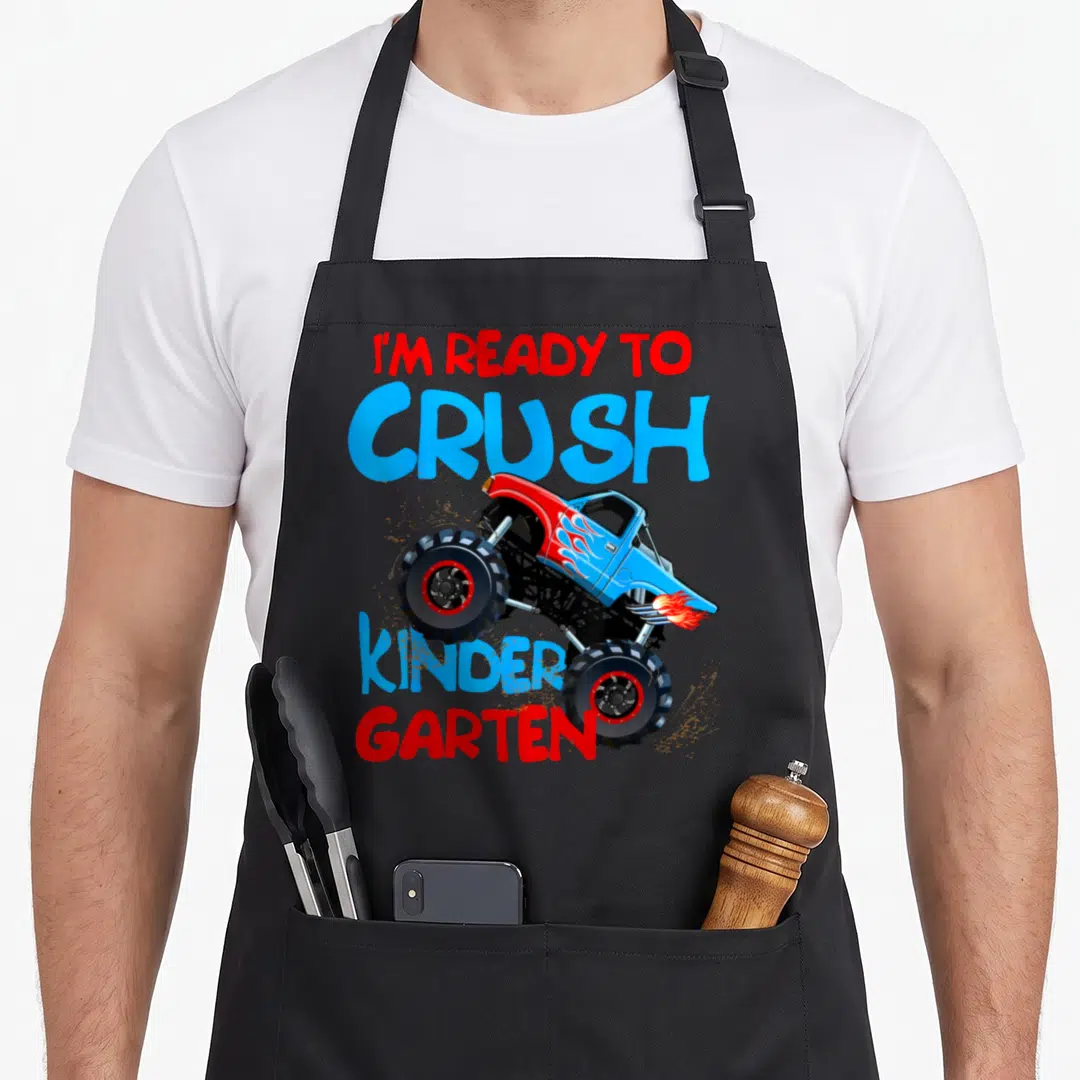School kindergarten truck apron - Monster truck graphic with I'M READY TO CRUSH KINDER GARTEN text in red and blue — shown on a black cotton canvas apron against a white background