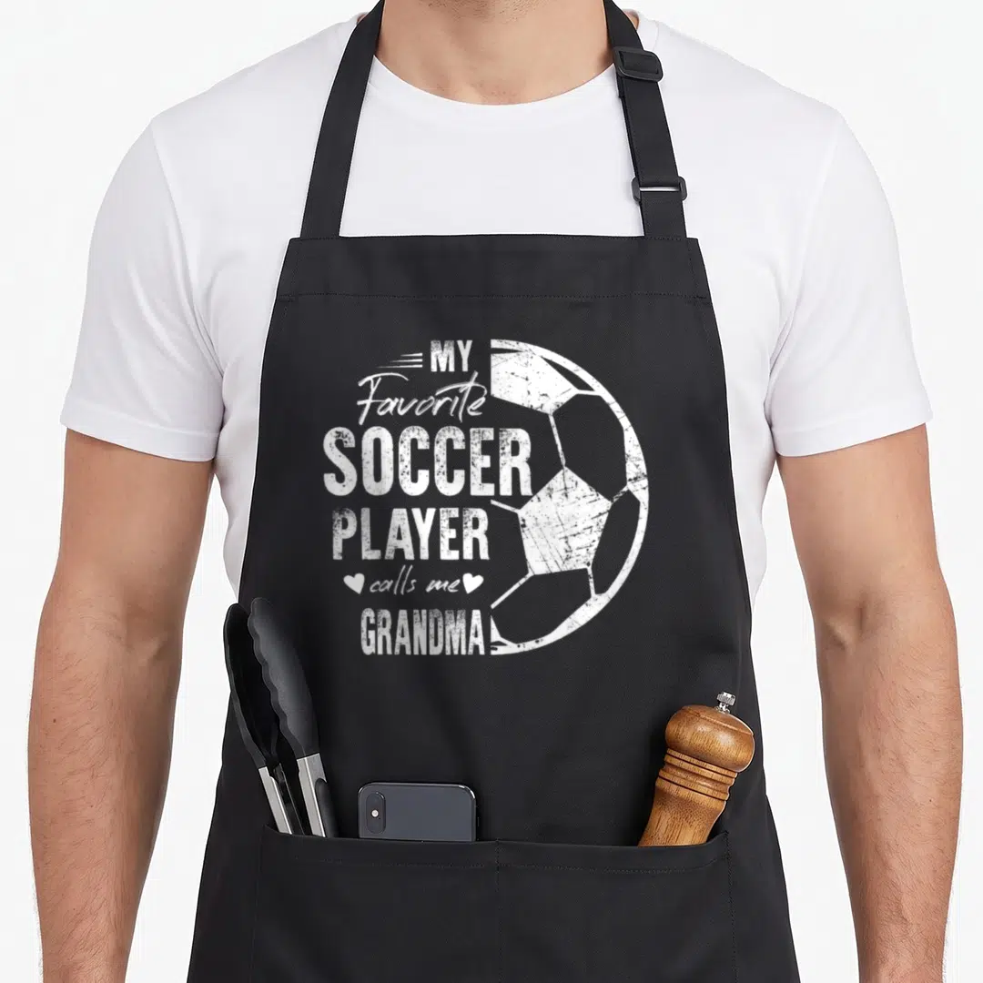 Player call grandma aprons - White distressed soccer ball and text MY Favorite SOCCER PLAYER calls me GRANDMA! with two — shown on a black cotton canvas apron against a white background