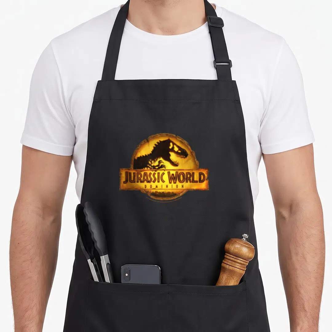 Dominion t-rex apron - Yellow circle with dinosaur silhouette and Jurassic World Dominion banner text — shown on a black cotton canvas apron against a white background