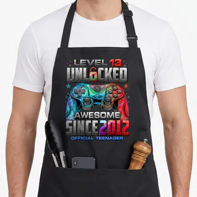 13th gaming aprons - Colorful game controller with Level 13 Unlocked text and Since 2012 lettering — shown on a black cotton canvas apron against a white background