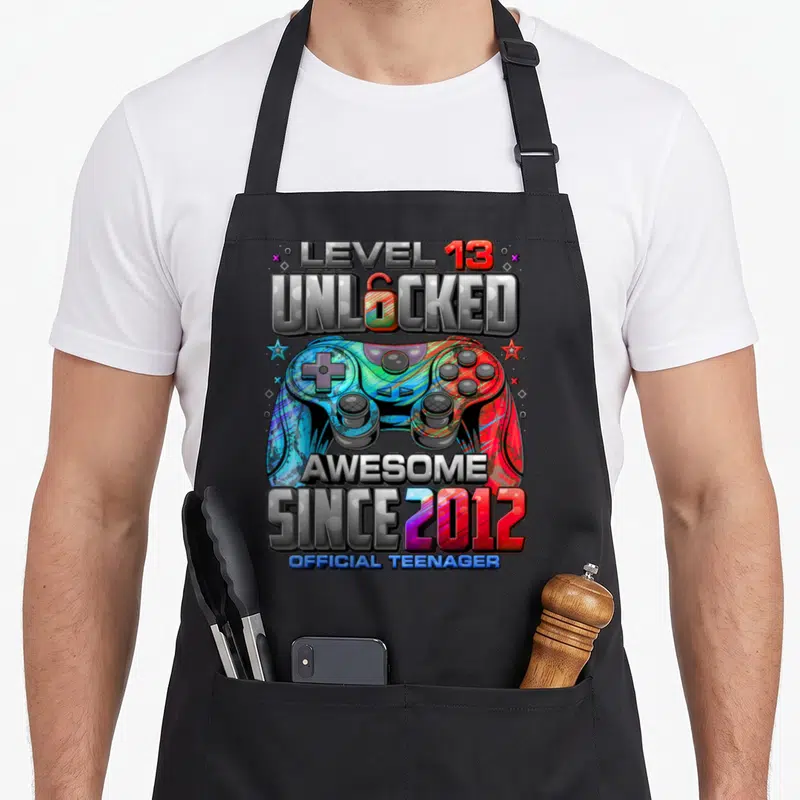 13th gaming aprons - Colorful game controller with Level 13 Unlocked text and Since 2012 lettering — shown on a black cotton canvas apron against a white background
