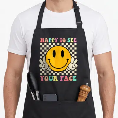 See teacher apron - Yellow smiley face with two peace sign hands and colorful text over a checkerboard pattern — shown on a black cotton canvas apron against a white background