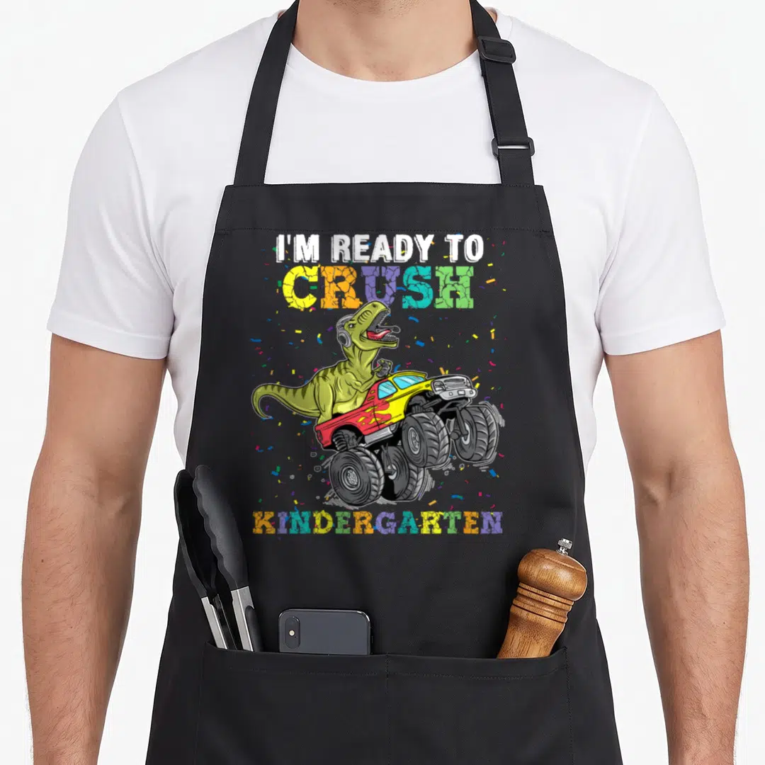 Ready truck apron - Dinosaur in monster truck with I'm Ready to Crush Kindergarten text and confetti — shown on a black cotton canvas apron against a white background
