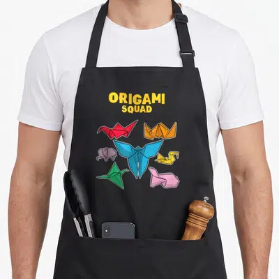 Squad paper apron - ORIGAMI SQUAD text above six colorful origami animals including a blue butterfly, red crane — shown on a black cotton canvas apron against a white background