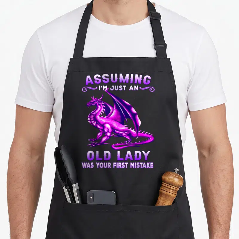 Mistake dragon apron - Purple dragon graphic between sarcastic text about being an old lady mistake — shown on a black cotton canvas apron against a white background