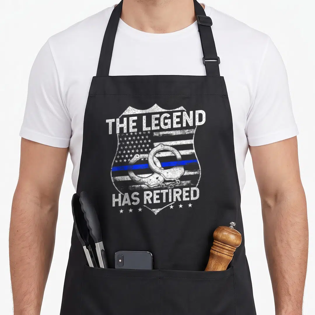 Police retirement apron - Distressed badge with handcuffs, thin blue line, and text THE LEGEND HAS RETIRED — shown on a black cotton canvas apron against a white background