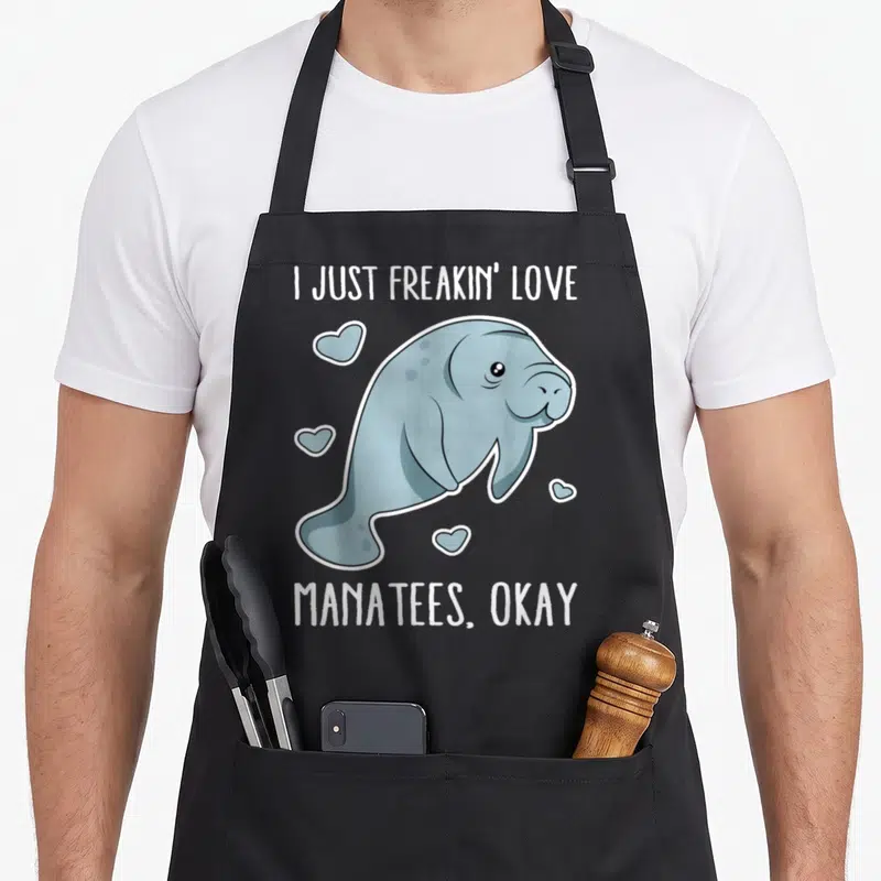 Freakin unique apron - Cartoon blue-grey manatee with white hearts and text I Just Freakin Love Manatees Okay — shown on a black cotton canvas apron against a white background