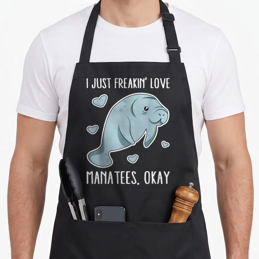 Freakin unique apron - Cartoon blue-grey manatee with white hearts and text I Just Freakin Love Manatees Okay — shown on a black cotton canvas apron against a white background