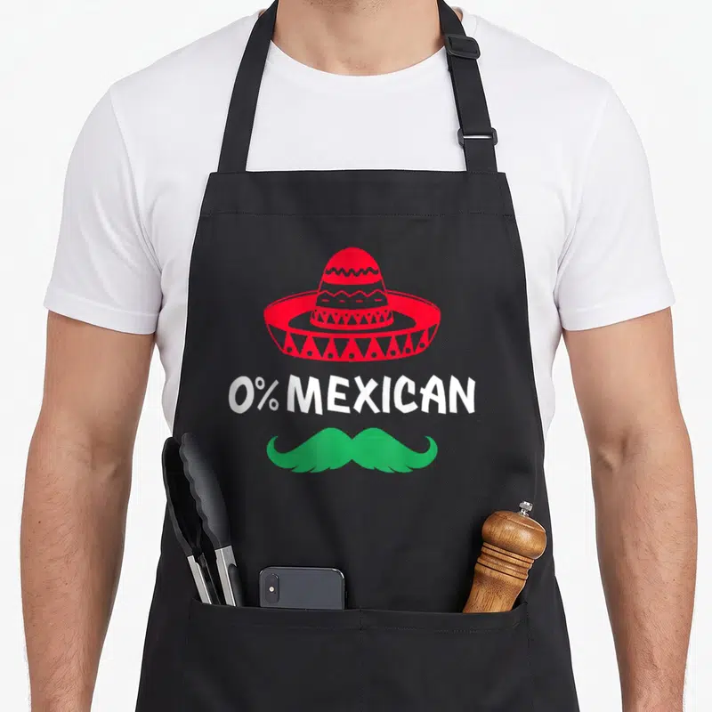 Mustache mexican apron - Red sombrero above white 0% MEXICAN text and a green handlebar mustache — shown on a black cotton canvas apron against a white background