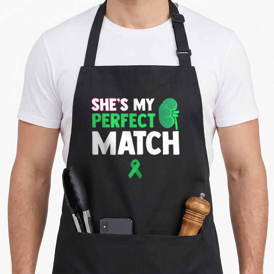 Recovering organ apron - SHE'S MY PERFECT MATCH text with cartoon green kidney and green ribbon — shown on a black cotton canvas apron against a white background