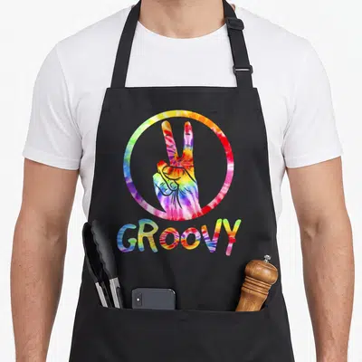 Groovy 70s tie-dye apron - Tie-dye hand making peace sign inside circle with groovy text below in red yellow green blue — shown on a black cotton canvas apron against a white background