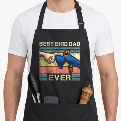 Father bird apron - Retro macav fist bumping human hand with BEST BIRD DAD EVER text on striped background — shown on a black cotton canvas apron against a white background